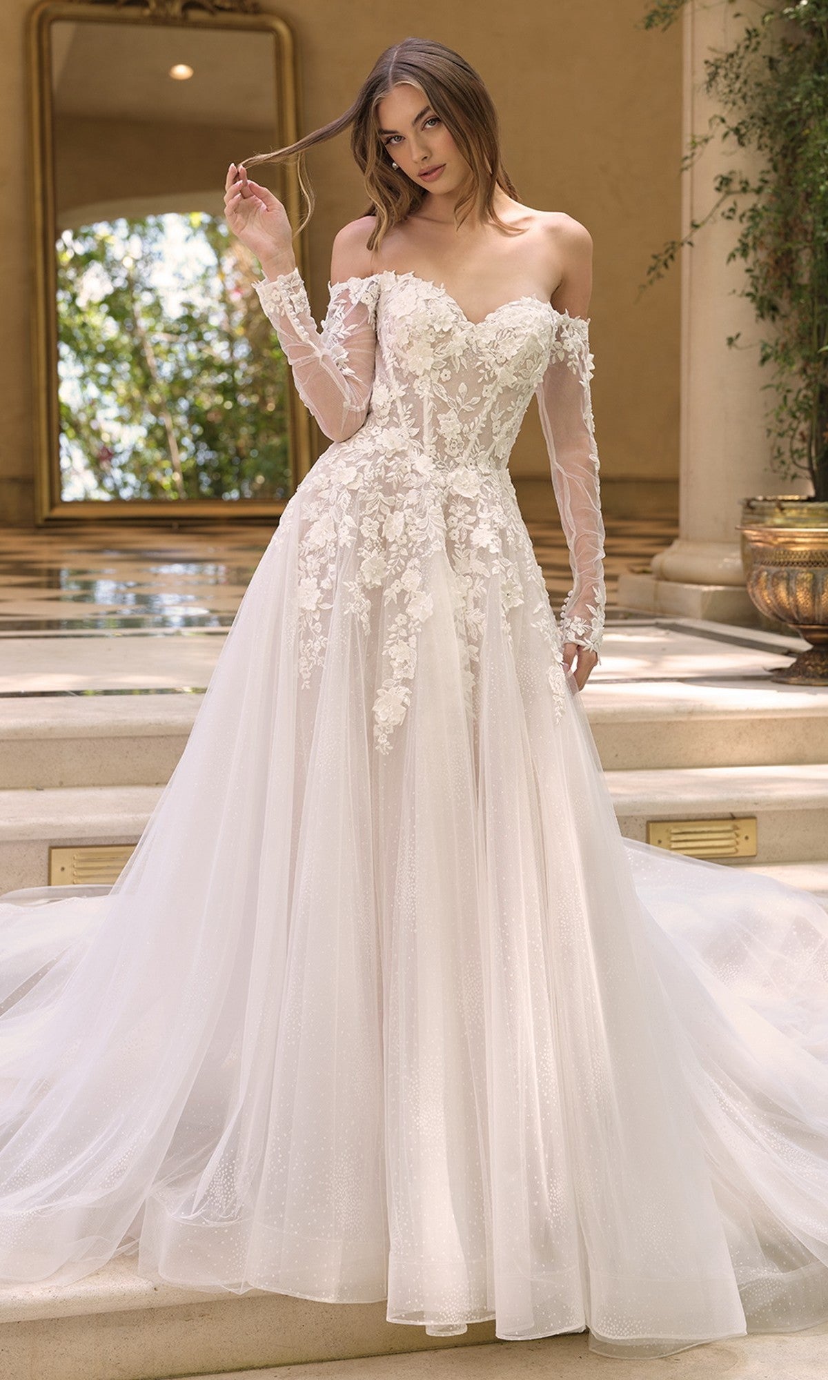 A woman stands on stone steps in the Andrea and Leo WL067 wedding dress, an off white/nude gown with a corset bodice, long sleeves, and a flowing tulle skirt detailed with 3D floral lace applique.